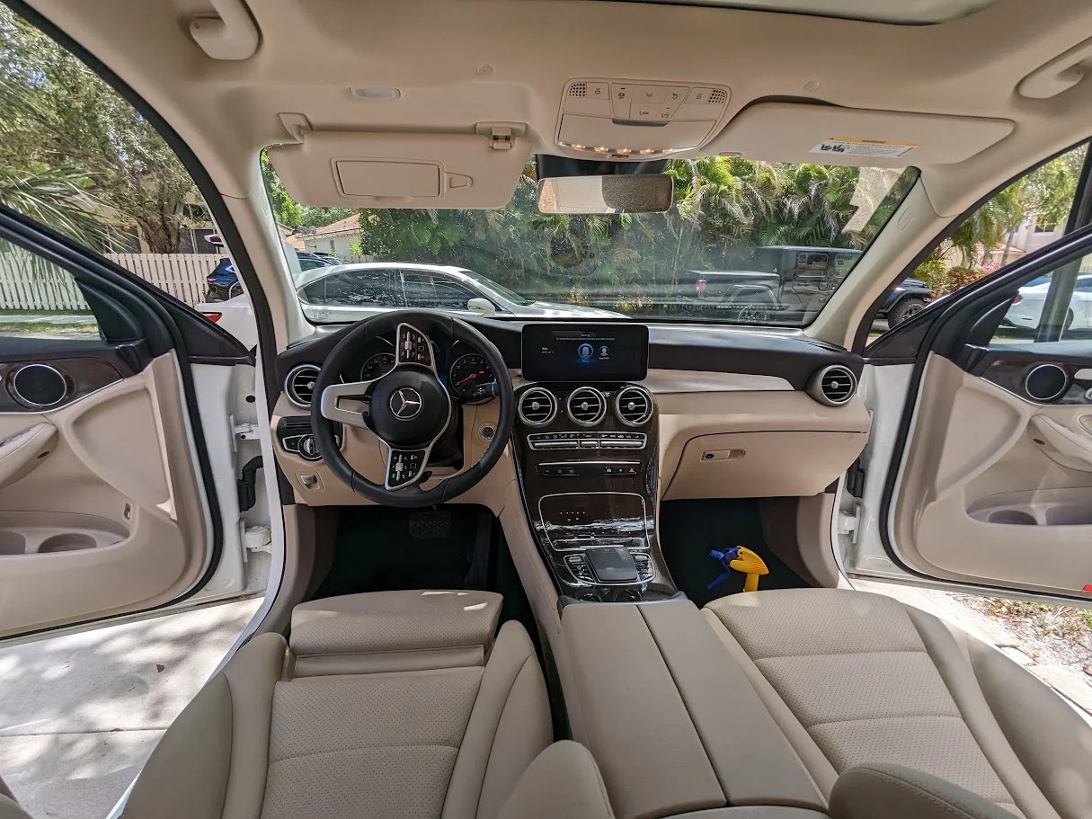 The interior of a white Mercedes-Benz vehicle is shown, with the doors open. The dashboard features a large infotainment screen, a steering wheel with the Mercedes-Benz logo, and wood-grain trim. The seats are upholstered in beige leather. Outside the car, a white fence, trees, and other vehicles are visible. A yellow spray bottle is on the floor of the passenger side.