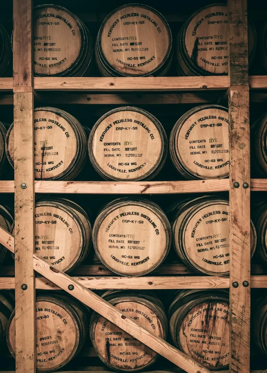 A detailed view of whiskey barrels stacked on wooden shelves in a distillery. The barrels are made of light brown wood and have dark lettering on their ends indicating the brand, contents, fill date, serial number, and location.