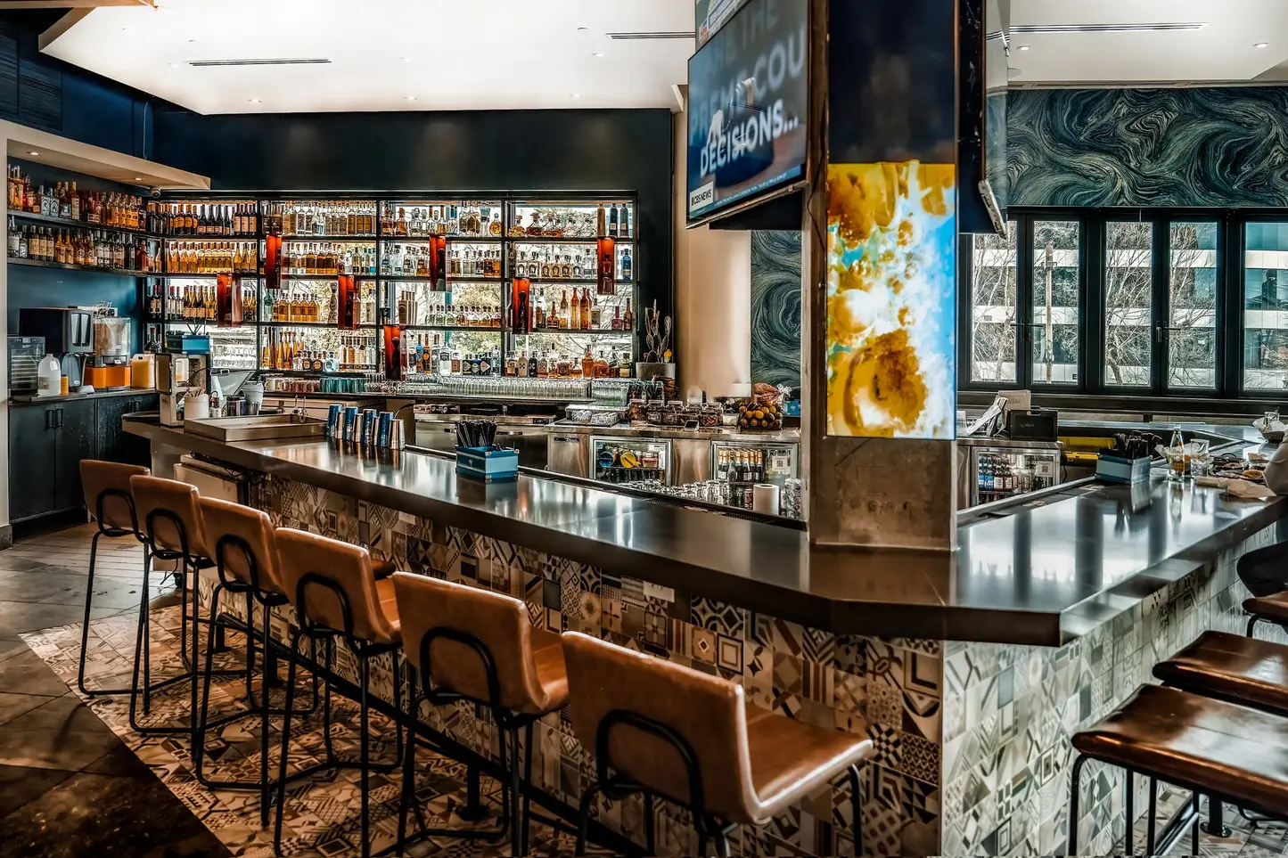 The bar area is vibrant and well-lit, with intricate tile patterns on the counter and a mirror-backed liquor shelf. It exudes a trendy and inviting atmosphere, perfect for social gatherings.