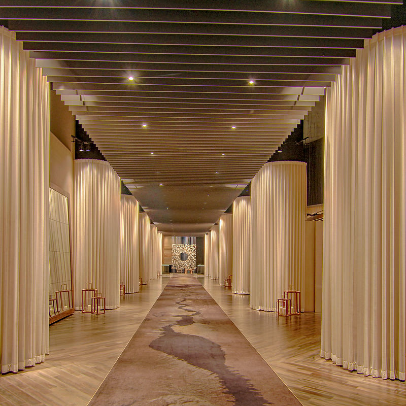 A long, wide hallway with cream-colored curtains draped along the sides and a patterned carpet runner down the center. The ceiling features a modern, slatted design with recessed lighting, and the floor is made of polished wood. At the far end of the hallway, a circular, textured art piece is mounted on the wall. Several small, wooden lanterns are placed along the sides of the hallway.