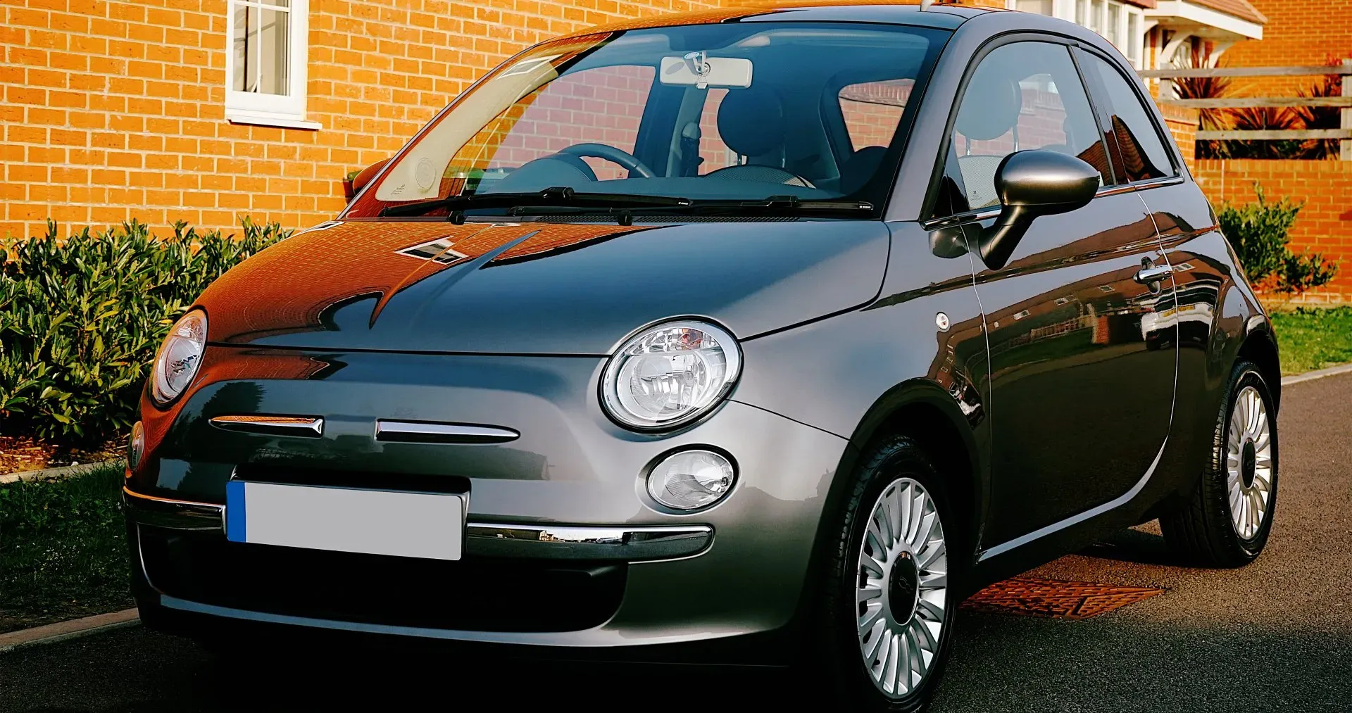 A glossy dark gray Fiat 500 is parked in front of a brick house with green bushes. The car is reflecting the surroundings, including the house and the sky. The vehicle is shown from a low-angle front-right perspective, highlighting its iconic design and polished finish. The license plate area is blurred out.