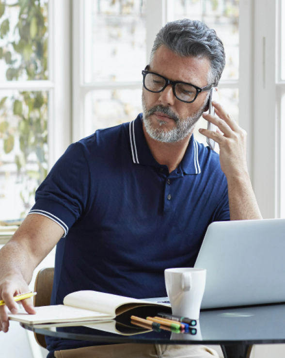 A middle-aged man with graying hair and a beard is sitting at a table, wearing glasses and a navy blue polo shirt. He is on the phone, holding a yellow pen in his left hand over an open notebook. A laptop, a white mug, and several colored pens are also on the table. He appears to be working or studying in a well-lit room with a window in the background.