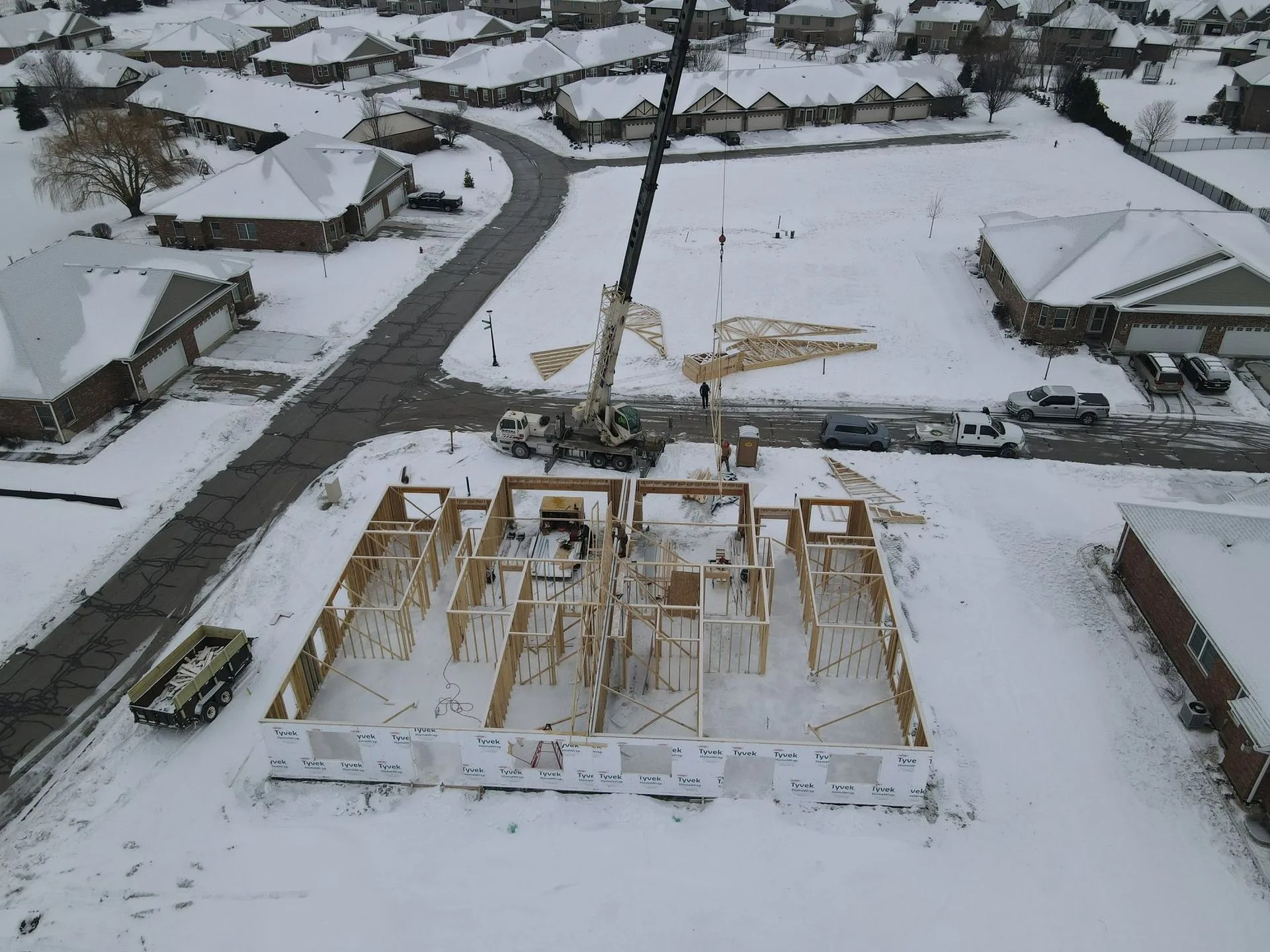 Aerial view captures residential home framing during winter construction, highlighting crane-assisted roof truss installation, professional builders, organized jobsite operations, structural framing progress, and efficient new home development.