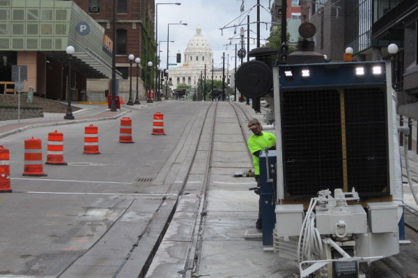 A vertical, mid-shot photograph capturing an operational specialty grinding machine on an urban street with embedded light rail tracks. The view is looking up a city hill. In the distance, centered perfectly, is the Minnesota State Capitol building with its prominent white dome. A DSI worker in a high-visibility green shirt stands near the controls of the white and black specialty grinder, which is operating between the rail lines.