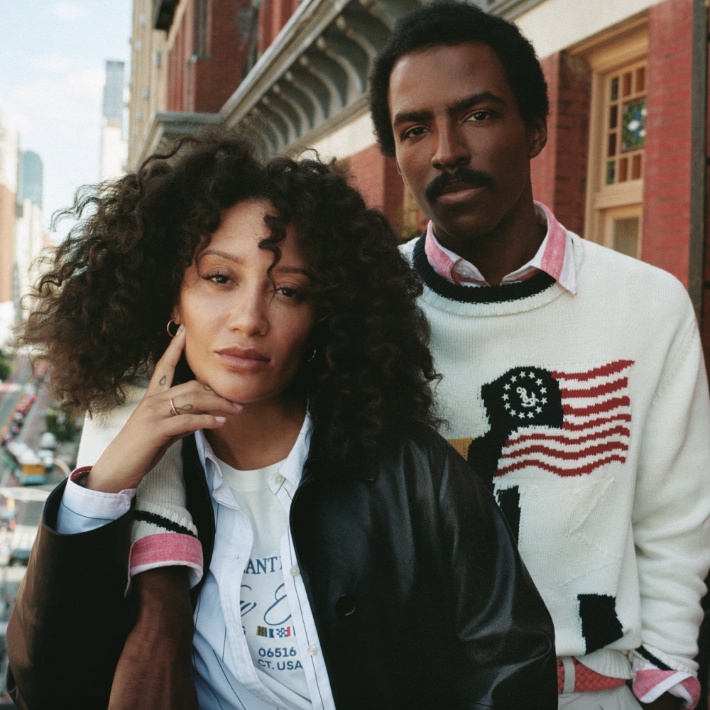 A stylish man and woman pose together on a city street. The woman has voluminous curly hair and wears a white graphic T-shirt layered under a blue button-up shirt and black leather jacket, with her hand resting against her chin. The man stands slightly behind her with his arm around her shoulder; he wears a white sweater featuring an American flag motif and a red collared shirt underneath. They stand in front of a red brick building with large windows.