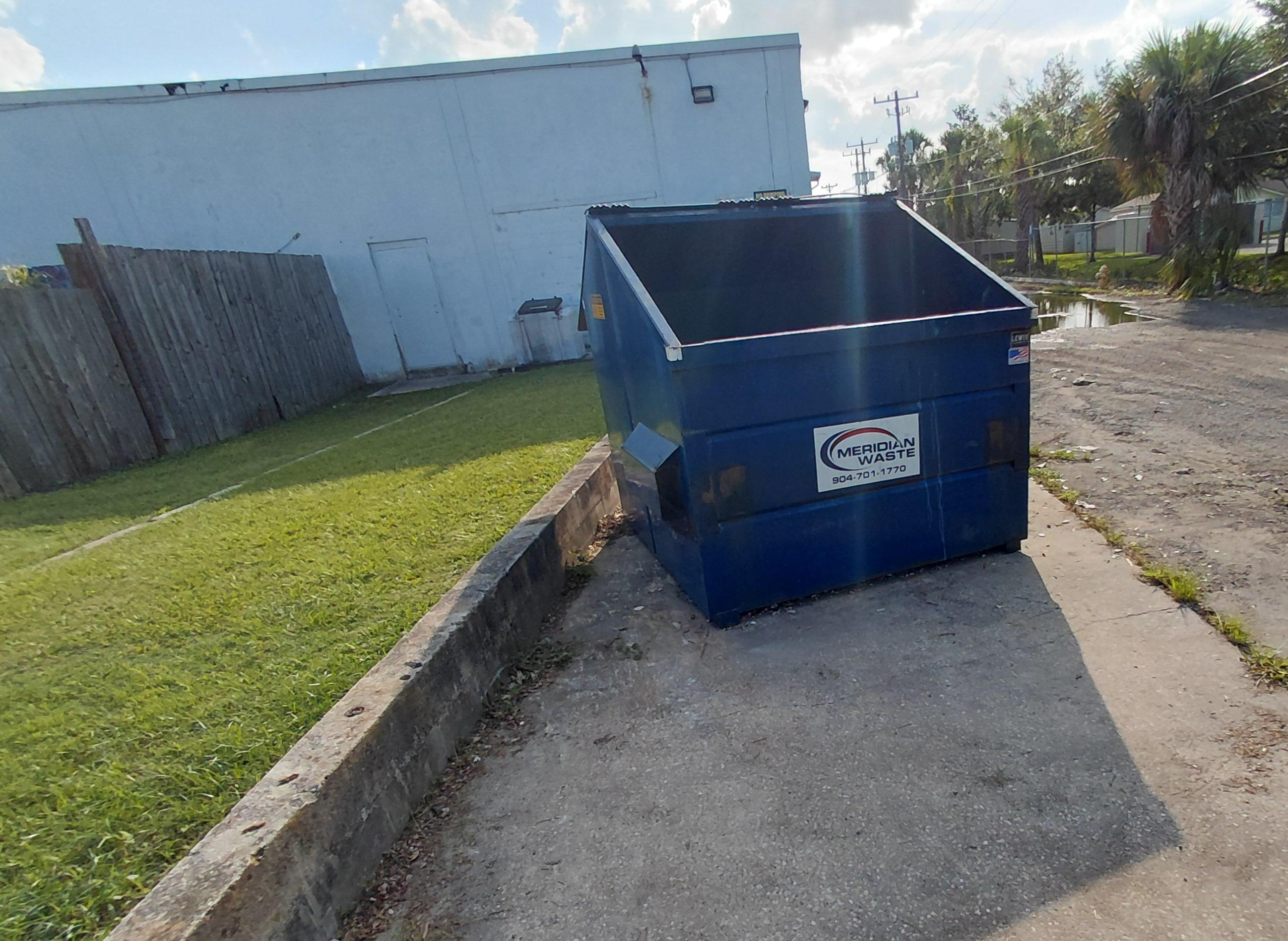 An empty blue commercial dumpster sitting on a clean concrete pad next to a lawn. 
