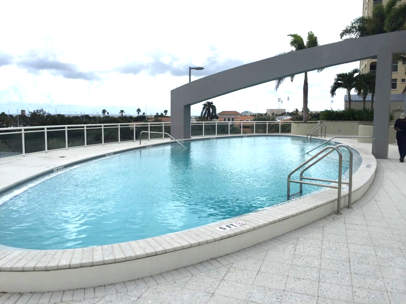 Large modern community swimming pool with clear blue water, curved edges, metal handrails, and an open pool deck.