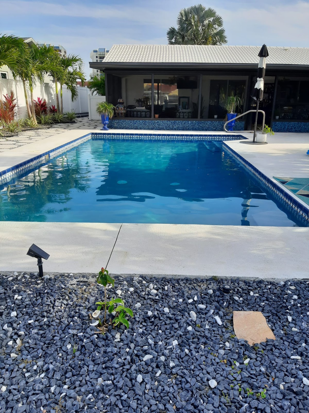 Rectangular backyard swimming pool with clear blue water, light stone coping, modern landscaping, and an outdoor living area.