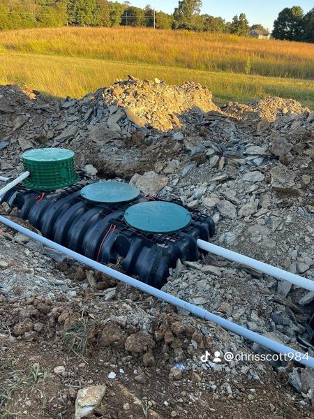 A black, corrugated septic tank with three green access lids is partially buried in a rocky excavation site. White PVC pipes are connected to the system against a field backdrop.