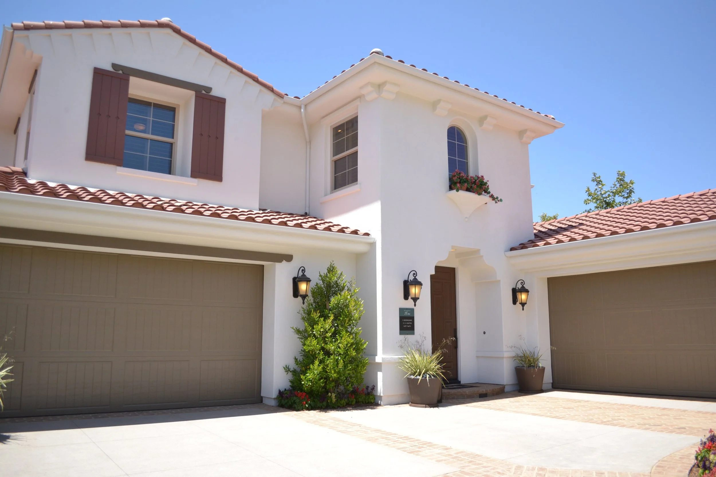 A large, white, two-story house with a tile roof and two garage doors. The house has brown shutters on one of the windows and a flower box above an arched window. There are outdoor lights on the front of the house and potted plants near the entrance. The sky is clear and blue.