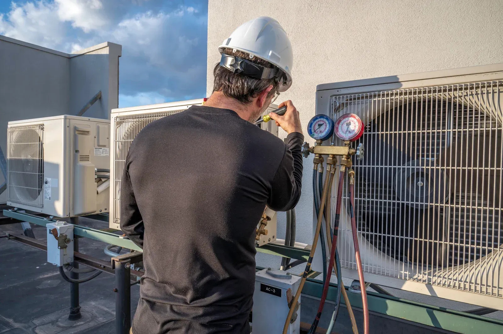 An HVAC technician wearing a white hard hat and goggles is on a rooftop, using a gauged manifold to service an outdoor air conditioning unit on a sunny day.
