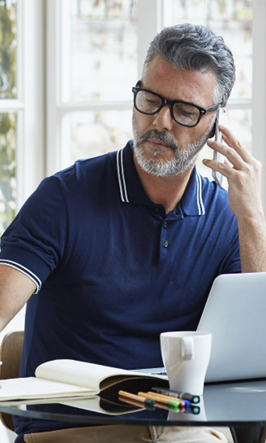 A middle-aged man with graying hair and a beard, wearing a navy blue polo shirt and black-rimmed glasses, is talking on a mobile phone while sitting at a desk. In front of him is a laptop, an open notebook, a white mug, and several colored pens.