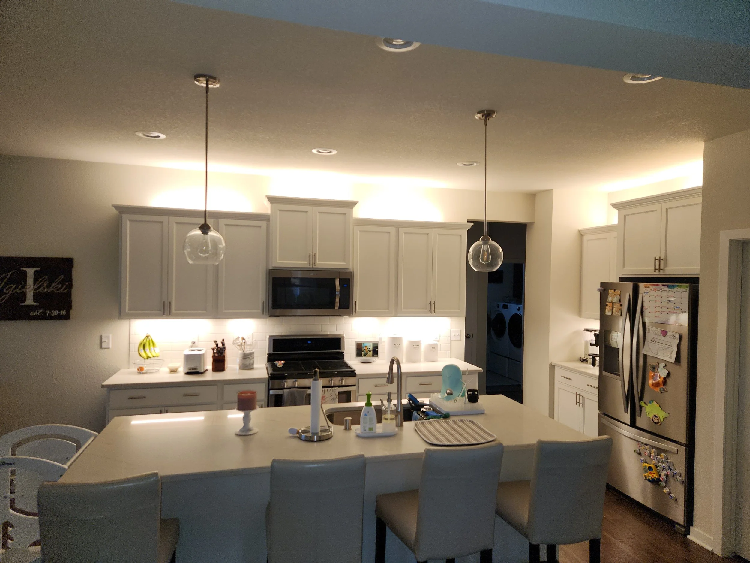 A well-lit modern kitchen with white cabinets, a central island with seating, and stainless steel appliances. The kitchen features recessed ceiling lights and pendant lights over the island. A large refrigerator is adorned with magnets and a calendar, and a wooden sign with a letter and date hangs on the wall. A baby high chair is visible on the left side of the island.