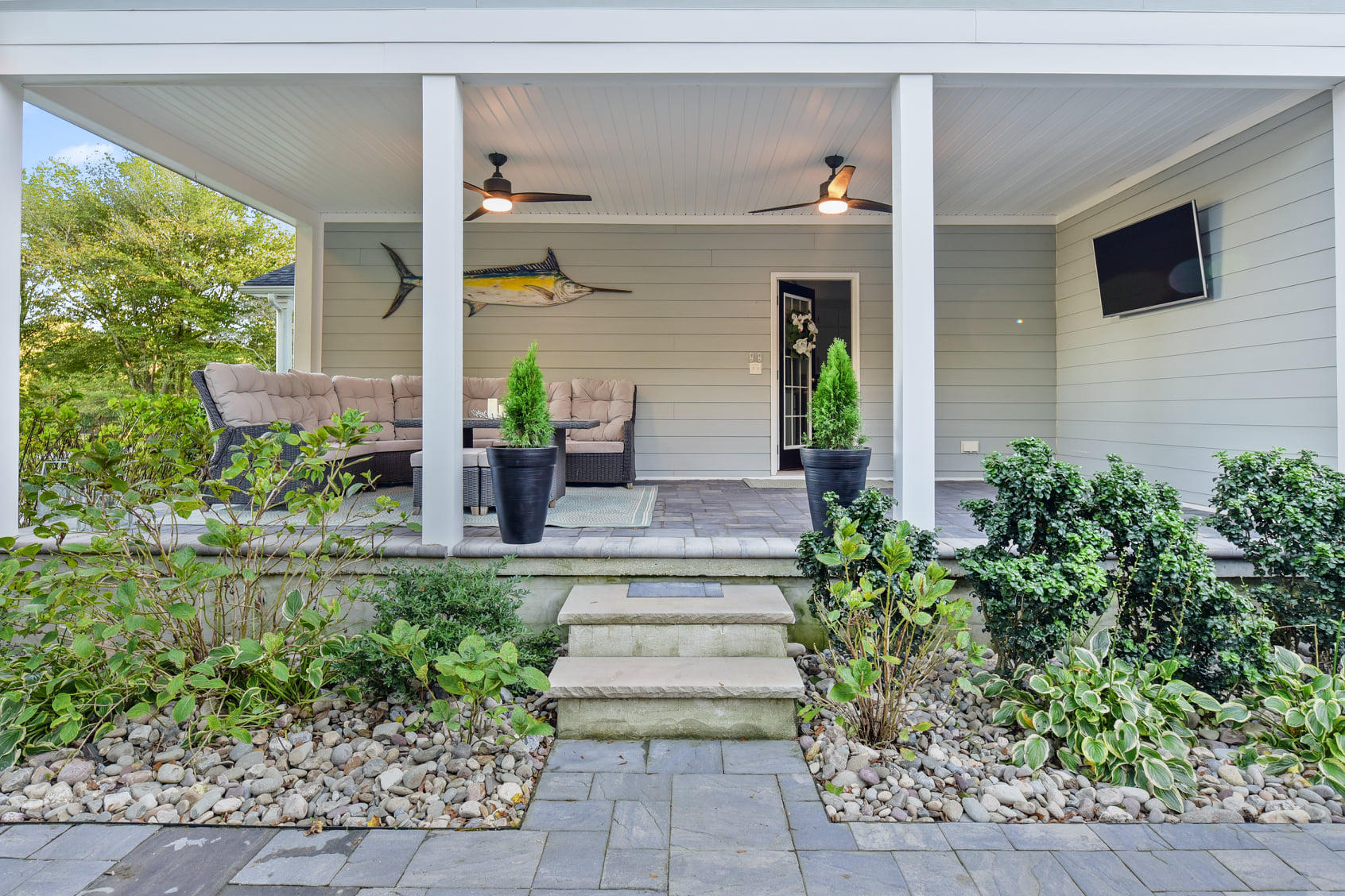 A professional hardscaping project underway in a Delaware home's backyard. The image shows landscapers laying irregular stone pavers to create a winding pathway. The scene includes a variety of landscaping tools and materials like sand and stone, with a partially completed patio area visible in the background. The house and garden display early spring foliage.