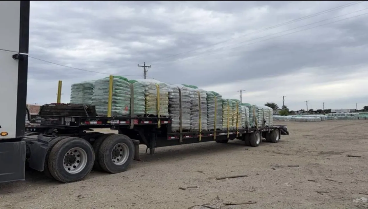 Flatbed semi truck carrying palletized bagged materials secured with straps in an industrial yard. Large shipment prepared for transport under cloudy skies, highlighting logistics, freight movement, and supply chain operations.