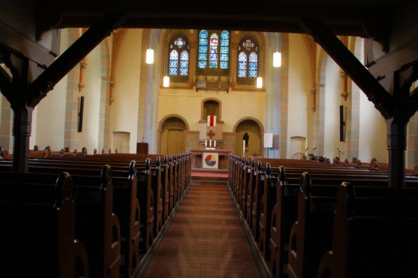 Unter der Empore angekommen öffnet sich der Blick auf den Kirchenraum, den Altar und den flachen Chorabschluss mit dem großen Drillingsfenster, dessen Originalverglasung aus der Bauzeit vom September 1899 bis Oktober 1900 erhalten geblieben ist. Auch die 