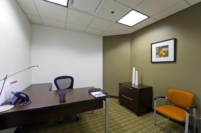 A compact and professional office space featuring a dark wooden desk with a sleek design, paired with a black mesh office chair. A bright orange guest chair adds a pop of color to the neutral-toned room, which includes olive-green and white walls for a calm yet modern aesthetic. A small cabinet with minimalist decor, including stacked white vases and framed abstract art, enhances the room's visual appeal.