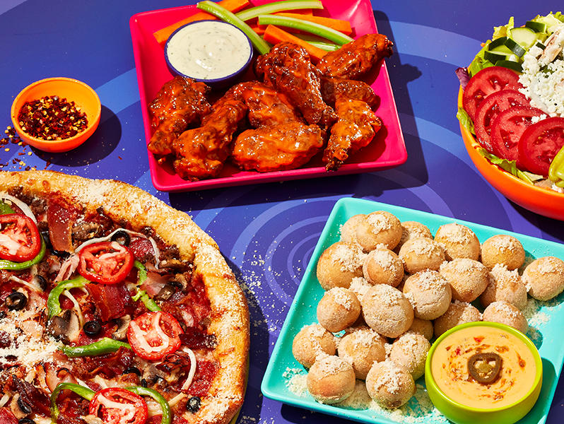 food spread featuring mellow mushroom pizza, pretzel bites, chicken wings, and salad