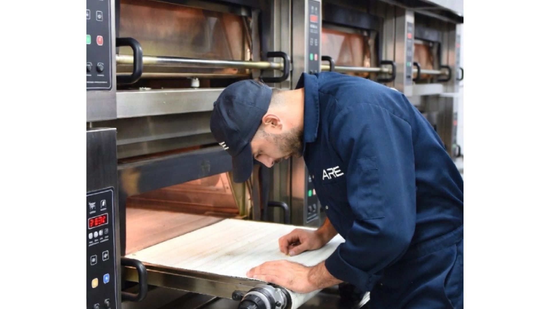 Professional technician in a navy ARE jumpsuit inspecting a high-volume industrial conveyor pizza oven for repair and maintenance.