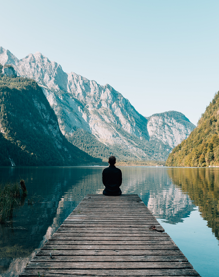 Person sitting at the edge of a dock, looking out at the calm lake and surrounding mountains.