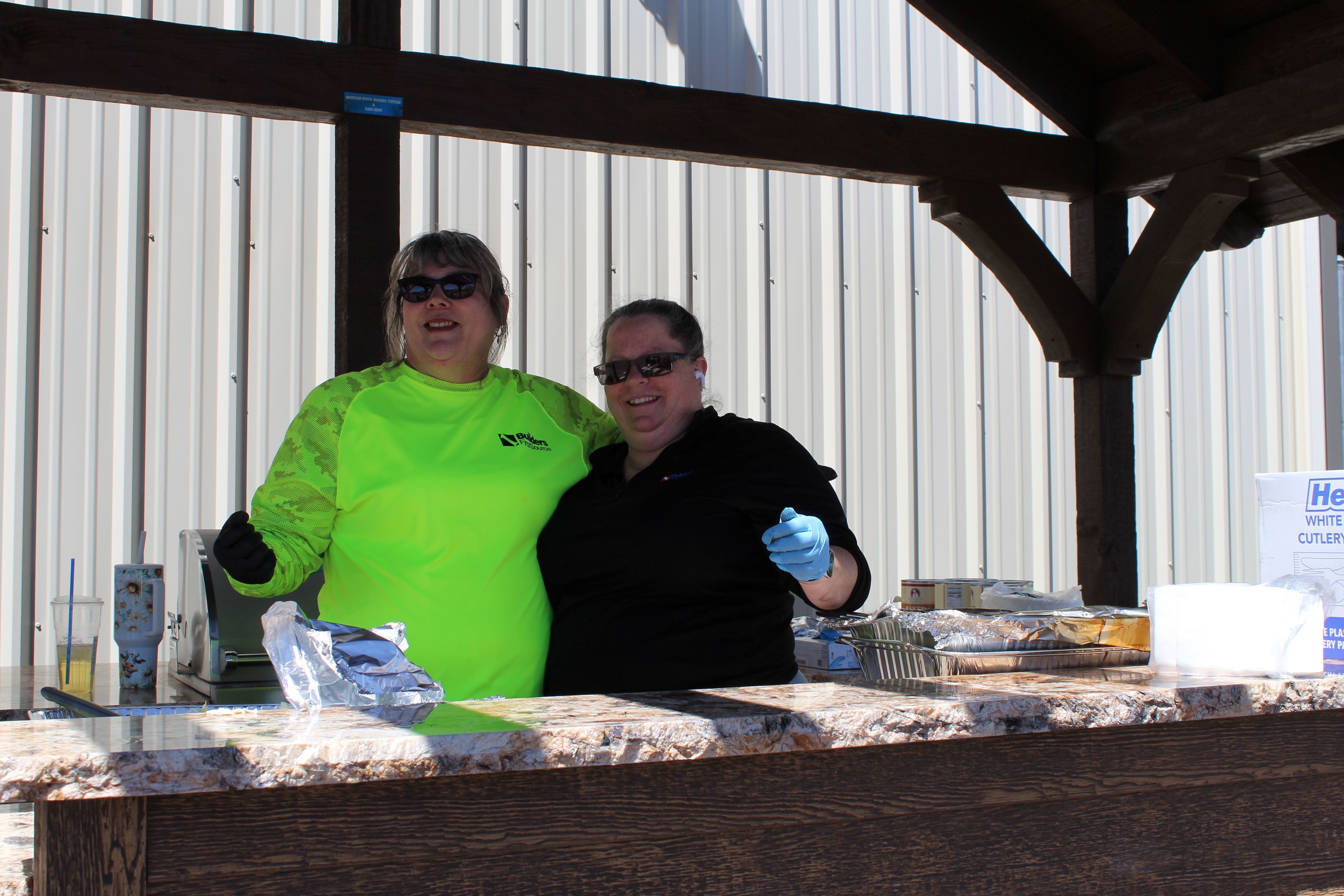 Two people standing behind an outdoor serving counter under a wooden shelter, wearing gloves and sunglasses, with foil-covered food trays and serving supplies arranged on the counter.