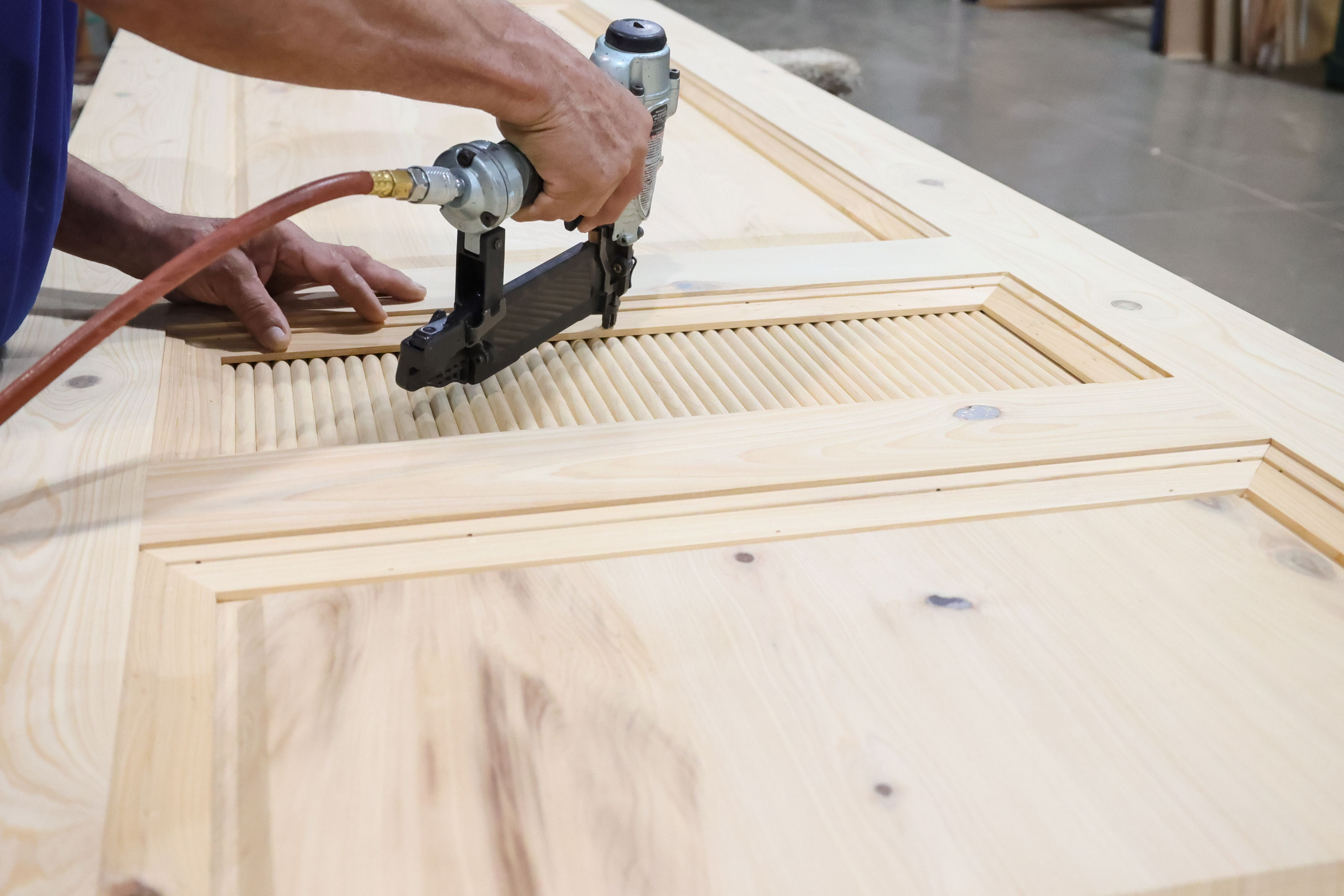 A close-up of a person’s hands using a pneumatic nail gun to assemble a wooden door. The door features a decorative louvered section in the center, and the wood grain is clearly visible. The worker is securing the pieces of the custom millwork door, which appears to be made of light-colored wood, possibly pine. The background is blurred, focusing attention on the detailed craftsmanship and tools being used.