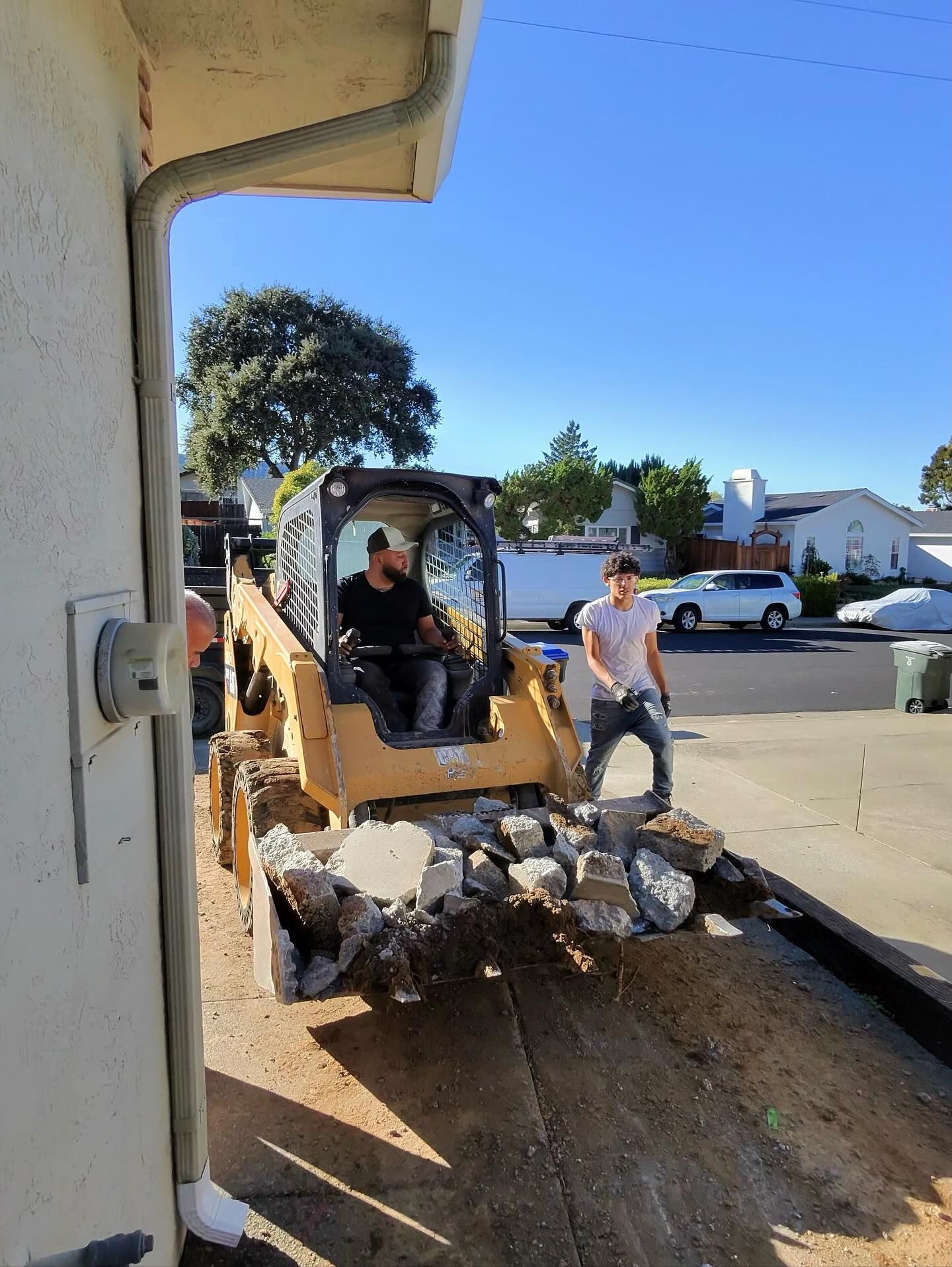 Construction workers removing broken concrete using a skid steer loader in a residential driveway, showcasing demolition, debris handling, and teamwork during site cleanup and preparation in a suburban neighborhood.