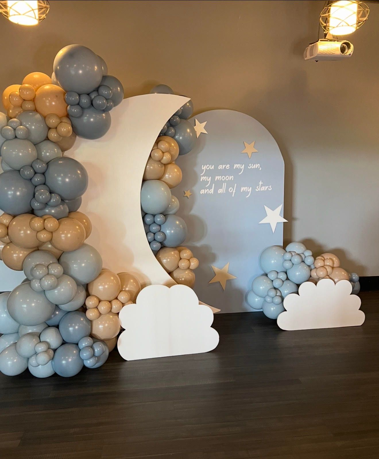 Dreamy baby event backdrop featuring crescent moon panel, soft blue and beige balloon garlands, cloud cutouts, star accents, and sentimental quote, creating a calm, celestial themed celebration setting indoors photo-ready.