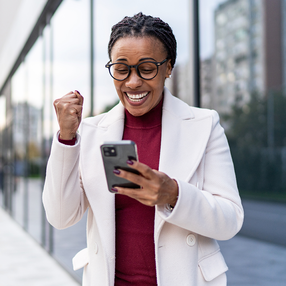 Person in a white coat holding a smartphone and making a fist pump gesture outdoors near a glass building.