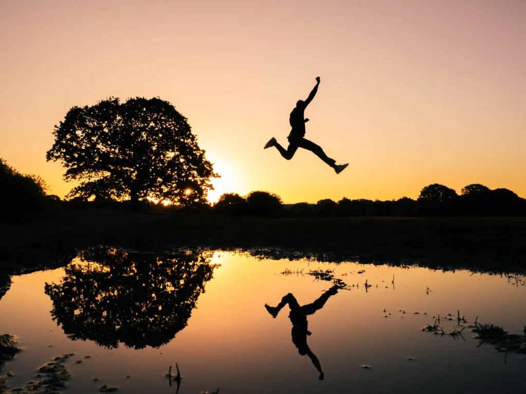 A silhouette of a person jumping in mid-air against a vibrant sunset sky, with a reflection visible in a pool of water below. A large tree is silhouetted to the left.