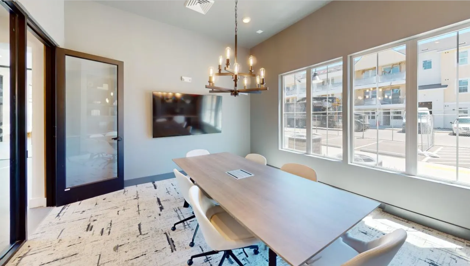 a conference room with a long wooden table, chic chandelier, and chairs surrounding with a TV and large, oversized windows