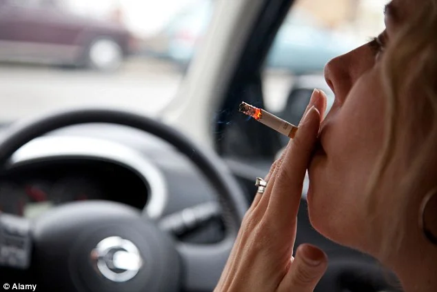 A close-up shot shows a woman's profile as she smokes a cigarette while driving a car. The steering wheel is visible in the foreground, and blurred cars can be seen outside the window. The image captures the act of smoking in a vehicle.