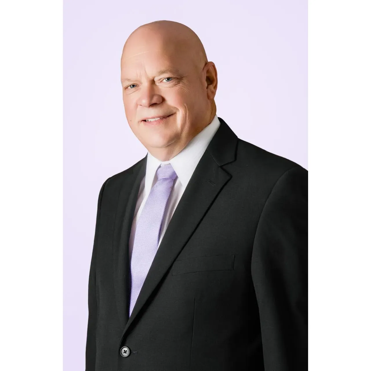 It&rsquo;s a professional studio-style headshot/portrait of a bald man wearing a black suit jacket, white dress shirt, and a light lavender/purple tie. He&rsquo;s angled slightly to the side, looking toward the camera with a small, friendly smile. The background is a very light lavender tone, giving the image a clean, polished, medical/professional look.