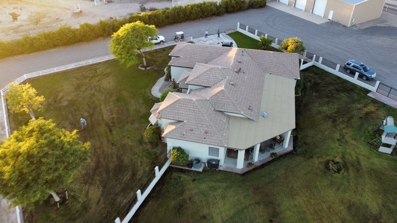 An aerial drone shot highlights a large, multi-gabled home with fresh tan tile roofing. The expansive property features a lush green lawn, white perimeter fencing, and mature trees during golden hour.
