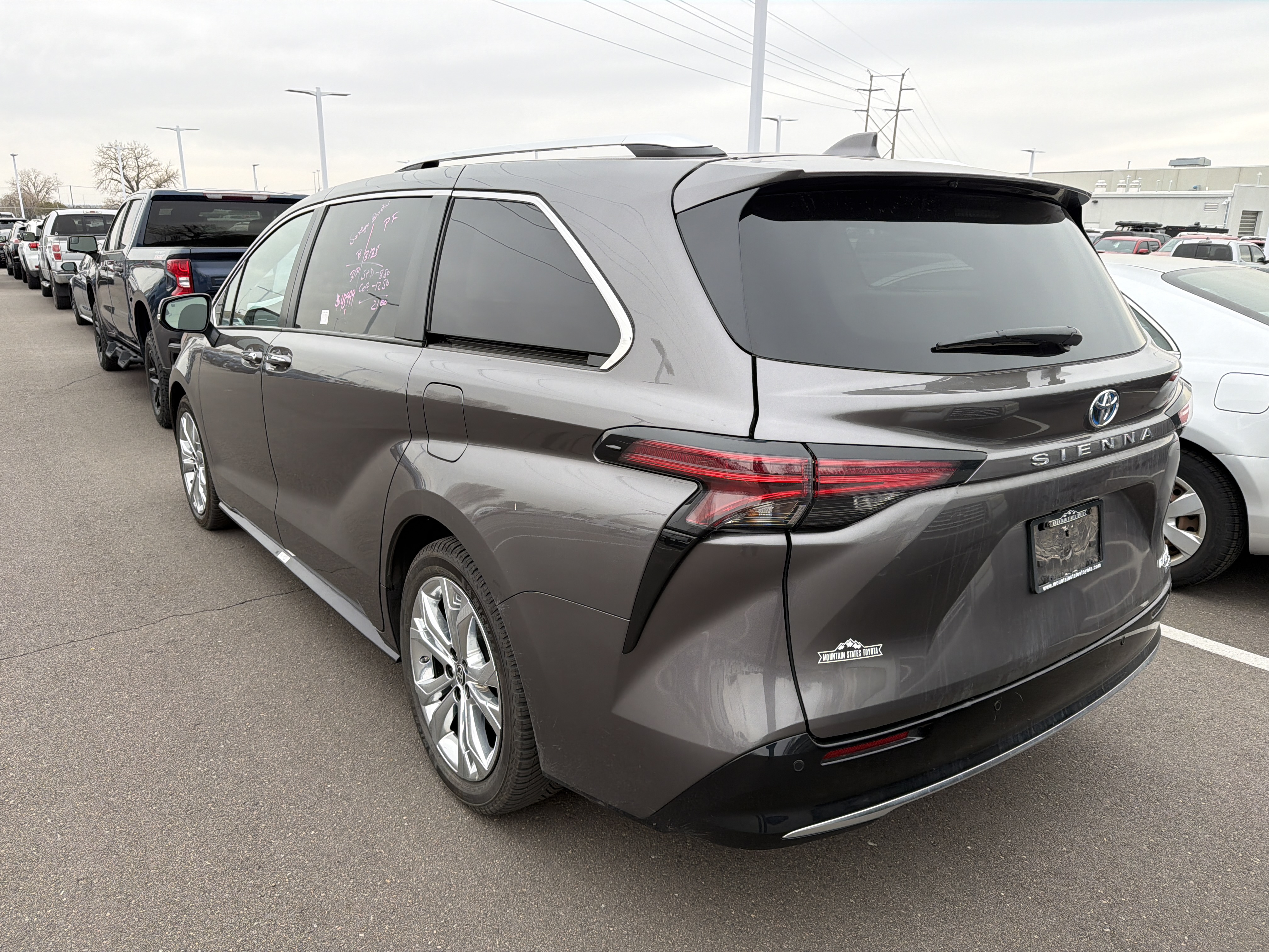 A dark gray 2023 Toyota Sienna minivan is parked in a lot with several other vehicles, including trucks and cars, under a cloudy sky. The minivan has a sticker on the rear passenger window with handwritten notes. A "Mountain States Toyota" sticker is on the rear bumper.