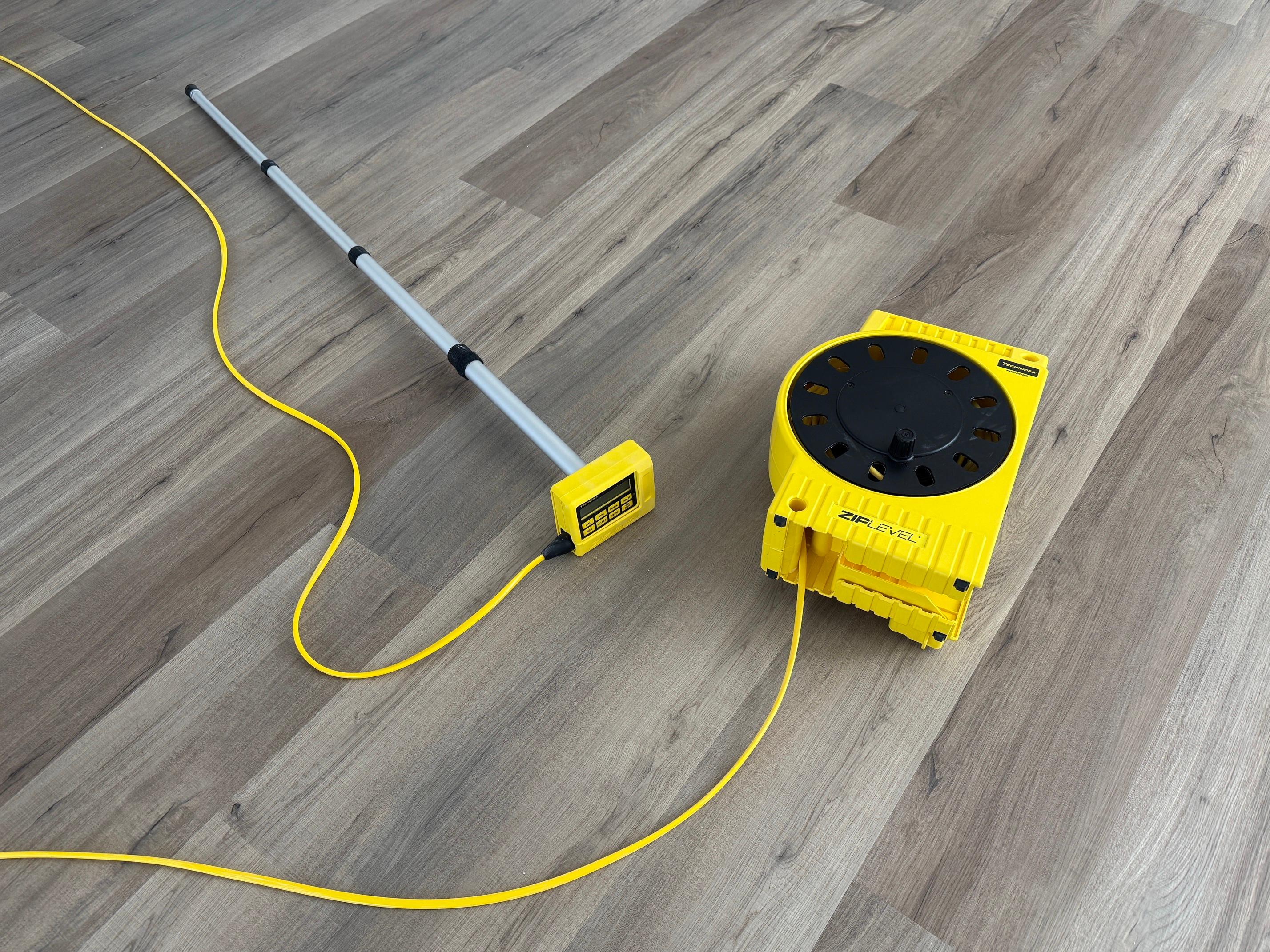 A yellow ZipLevel Pro elevation measurement tool with its probe and cable laid out on a gray vinyl plank floor.