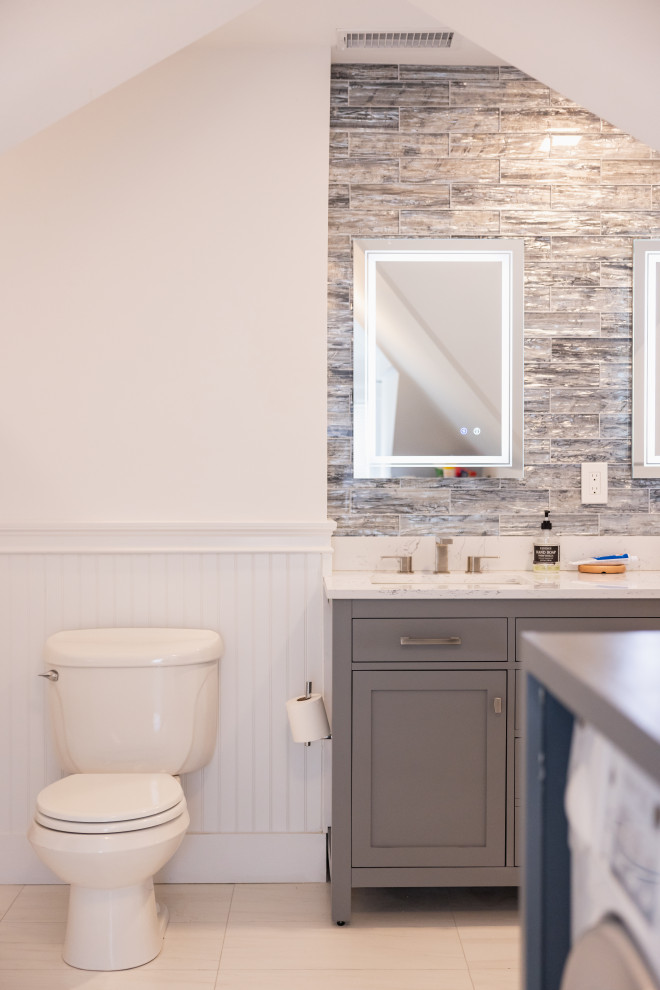 A well-lit bathroom features a modern vanity with a grey cabinet, a white countertop, and a sink with silver faucets. Above the vanity, a rectangular mirror with integrated LED lighting is mounted on a wall tiled with grey and brown rectangular tiles. To the left of the vanity, a white toilet is positioned against a white beadboard wall. A toilet paper holder with a roll of toilet paper is attached to the wall next to the toilet. The floor is tiled with light-colored square tiles. In the foreground, a portion of a blue washing machine is visible.