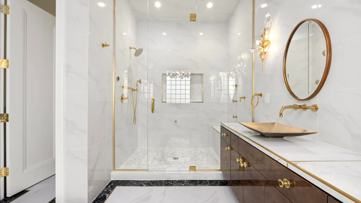 A luxurious bathroom with marble tile walls, a glass-enclosed shower with gold fixtures, and a vanity with a gold vessel sink and dark wood cabinets. A round gold-framed mirror hangs above the vanity.