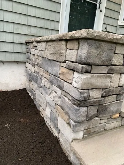A stone-clad column next to a house with green siding and a white door. The column is constructed from irregular-shaped, stacked stones in various shades of gray and tan. The ground to the left of the column is covered in dark soil, suggesting a garden bed or landscaping. To the right, a concrete step or landing is visible.