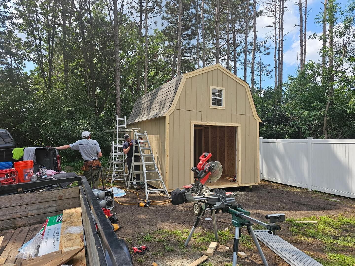 Two workers are building a gambrel-roofed storage shed with tan vertical siding in a wooded area. Construction tools, including a miter saw, are set up in the foreground, and a white vinyl fence is visible to the right.