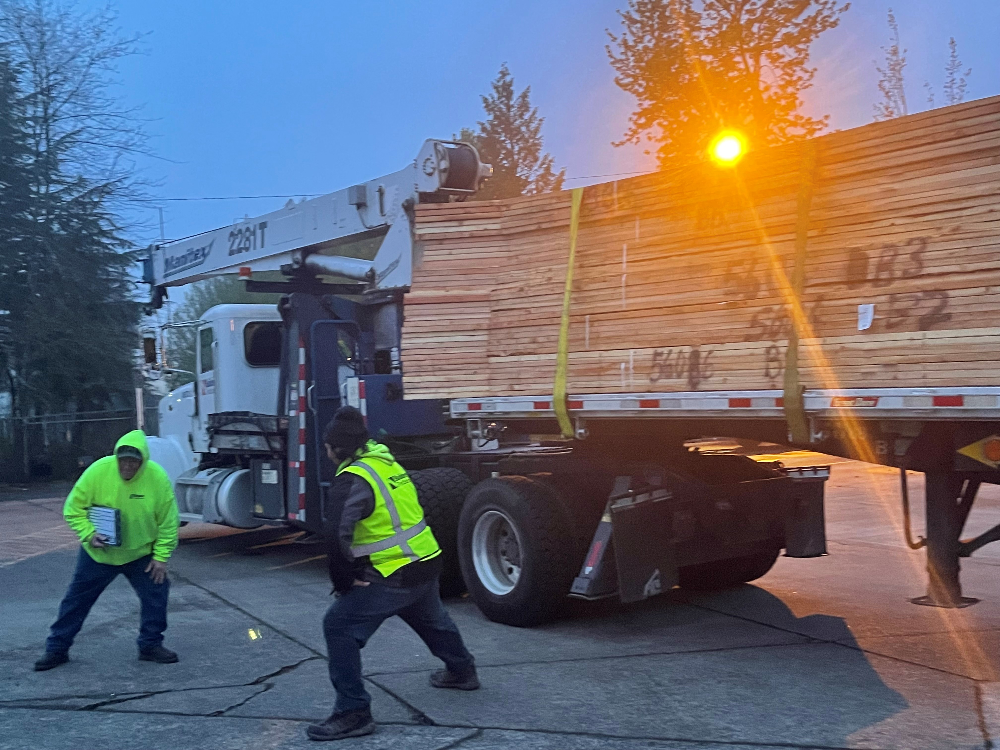 Two workers in high-visibility safety gear posing in front of a flatbed truck. The truck is loaded with a large stack of bundled lumber, and one worker is holding a clipboard. It is early morning with a slight blue hue in the sky with the sun rising above the lumber truck.