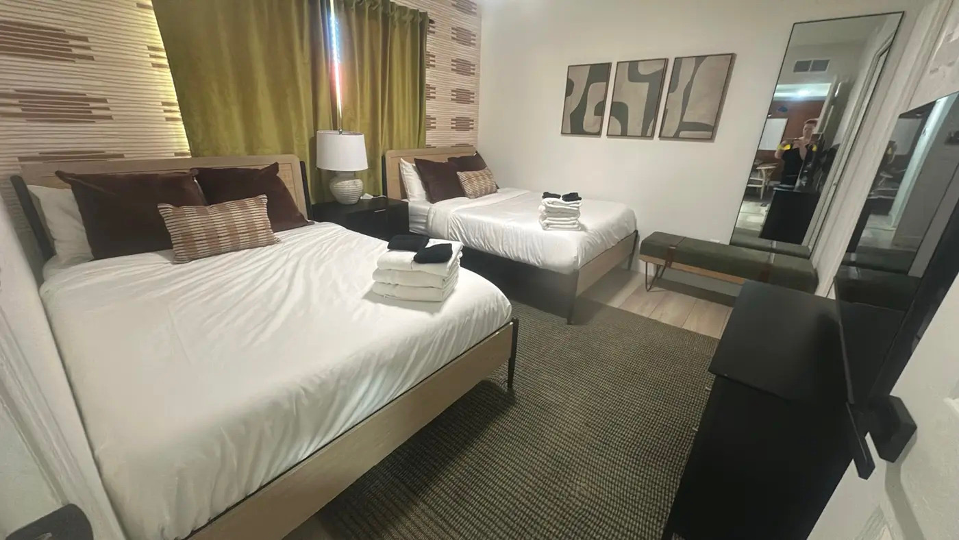 A staged guest bedroom with two beds, neatly made with white linens and brown pillows. Stacks of folded towels and dark washcloths are placed on each bed. The room features modern decor, including abstract art on the wall, a full-length mirror, and a dark dresser. A green velvet bench sits near the mirror. The overall aesthetic is clean, neutral, and designed for a vacation rental.