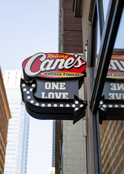 A vertical, projecting outdoor sign attached to a building corner featuring the red Raising Cane's logo above a black marquee-style arrow reading "ONE LOVE," set against a backdrop of tall city skyscrapers.