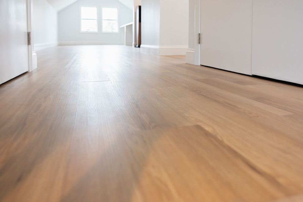 A low-angle shot of a modern, empty room featuring light-colored hardwood floors and white walls and doors. Two windows are visible in the background, letting in natural light that reflects off the polished floor. The overall impression is one of clean, minimalist design.