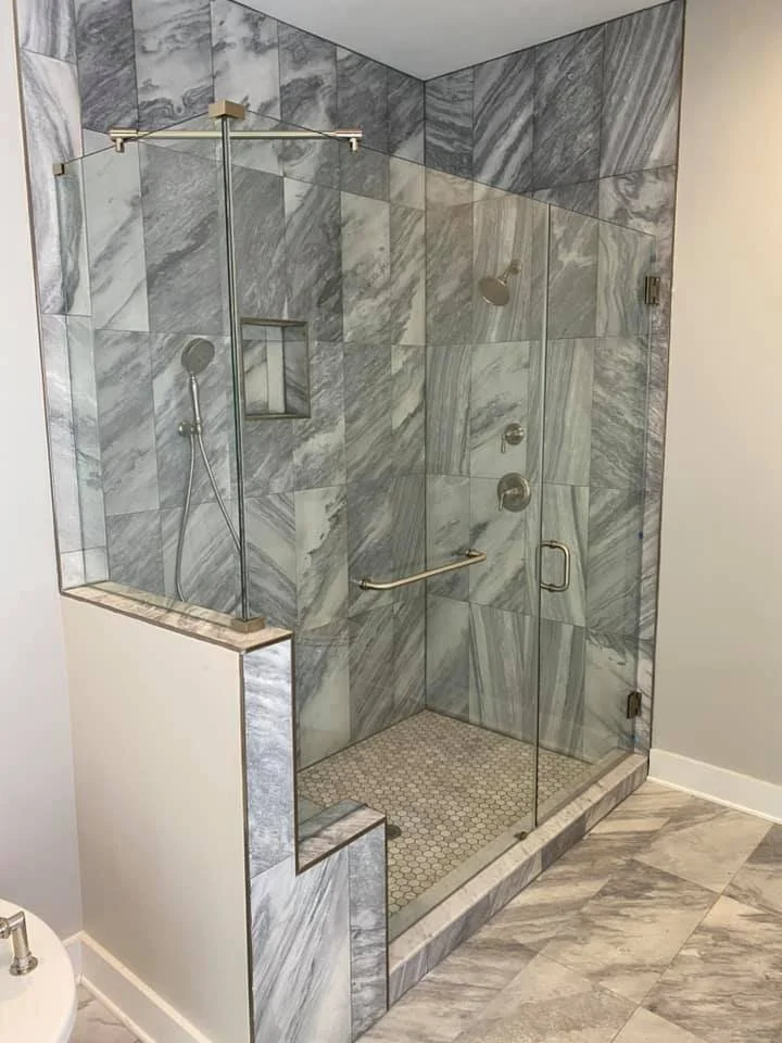 A modern bathroom features a spacious glass-enclosed shower with marble tile walls and a hexagonal tile floor. The shower has brushed nickel fixtures, including a rainfall showerhead and a handheld showerhead, as well as a long horizontal handle on the glass door. The surrounding bathroom floor is also tiled with a marble pattern. A portion of a white toilet is visible in the lower left corner.
