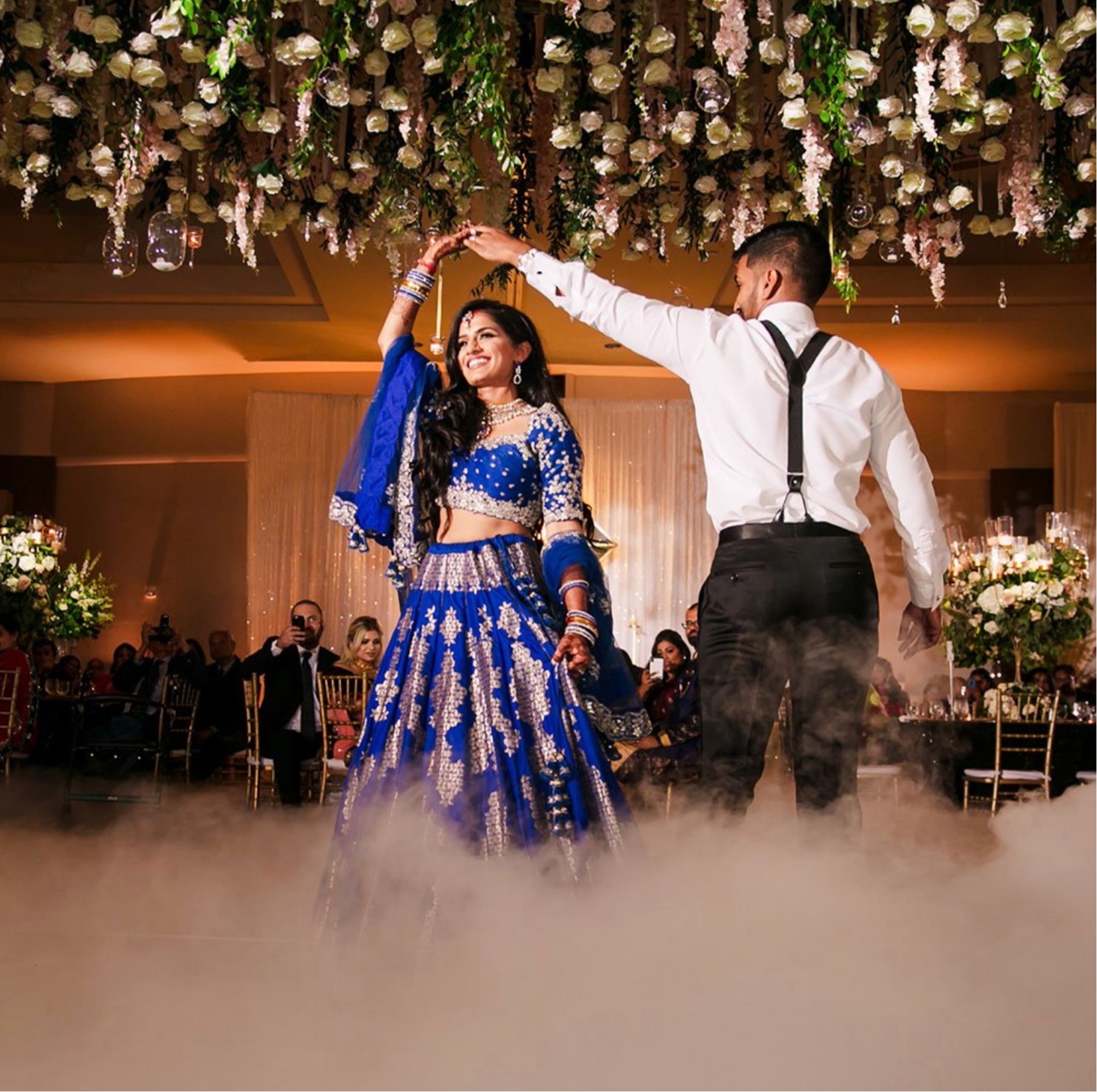 A vibrant celebration brought to life by WS Event Production Inc.'s stunning lighting and décor. The elegant overhead floral installation, accented by soft golden lighting, creates a dreamy canopy for the couple's dance. Low-lying fog adds a magical touch, drawing focus to the couple while enhancing the luxurious ambiance of the space. The lighting perfectly highlights the rich blue and silver tones of the bride's attire and the lush floral arrangements, delivering an unforgettable experience for all in attendance.