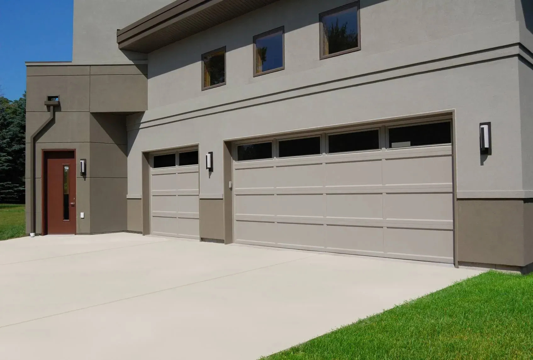 A modern home showcases two white sectional garage doors with narrow upper windows, clean architectural lines, neutral walls, contemporary lighting, and a spacious concrete driveway with polished curb appeal today.