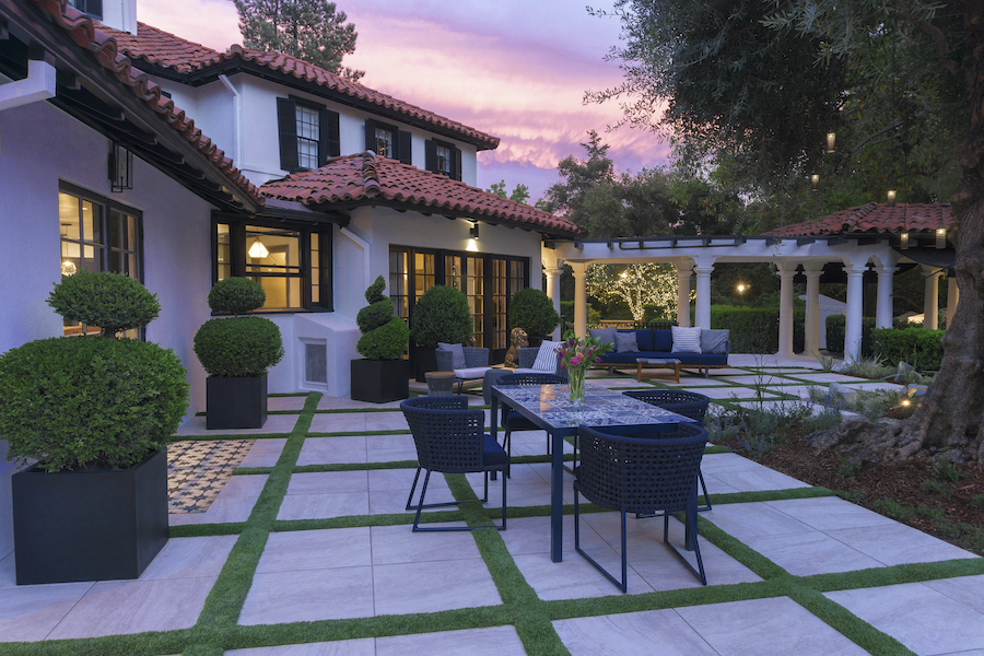 Spanish-style courtyard at sunset, featuring a tiled villa, manicured topiary, outdoor dining area, white columns, and warm garden lights.