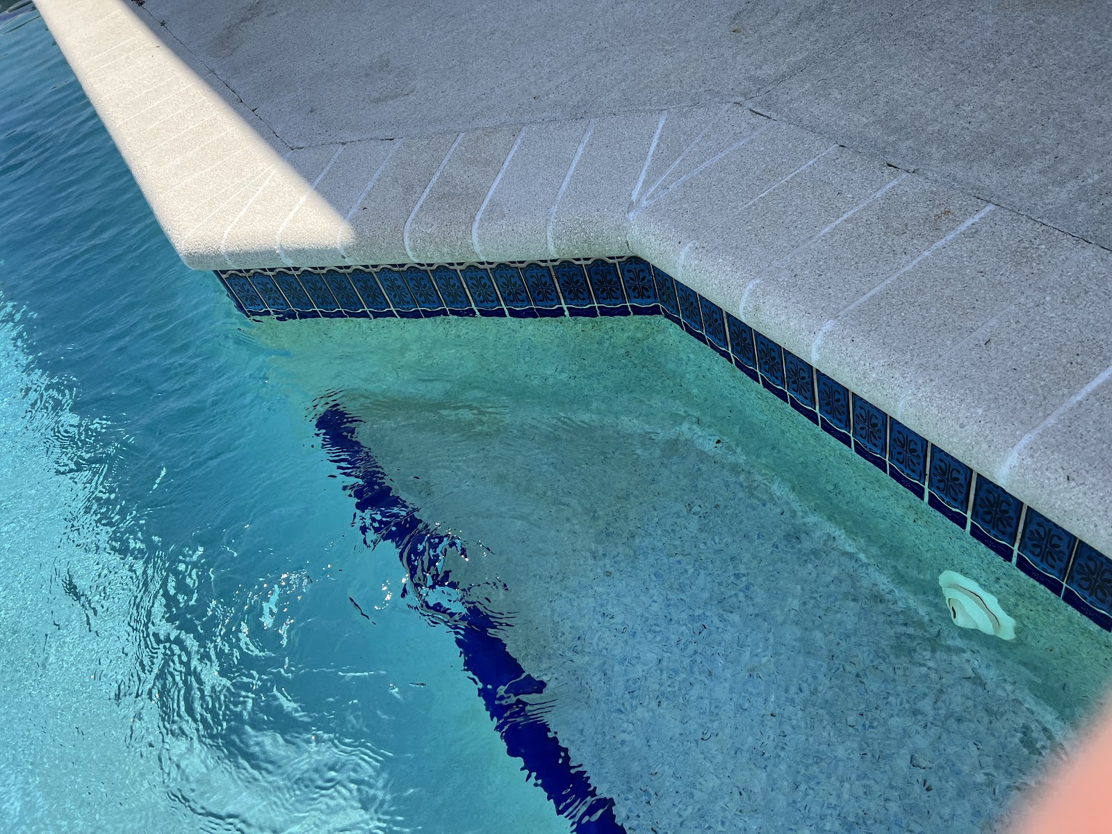 Close-up of a residential swimming pool corner with clear blue water, decorative tile, and light stone coping.