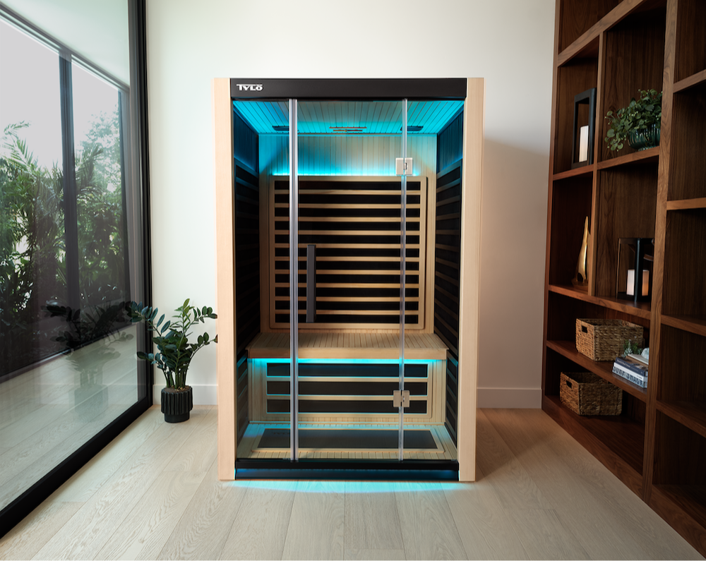 A Tyl&ouml; Kiruna 2 infrared sauna is shown as a front-facing product lifestyle shot in a bright, modern residential setting. The unit features a light aspen wood frame with a black interior, full-length glass double doors with chrome handles, and striking teal/aqua chromotherapy LED lighting along the ceiling and floor perimeter. The interior reveals slatted infrared heating panels along the back wall and a two-tiered bench system with a natural wood floor grate. The Tyl&ouml; logo is prominently displayed on the black top trim. The sauna is staged in a stylish living space with light hardwood floors, a large floor-to-ceiling window, tropical potted plants, and a dark walnut open bookshelf, conveying a luxury home wellness aesthetic.